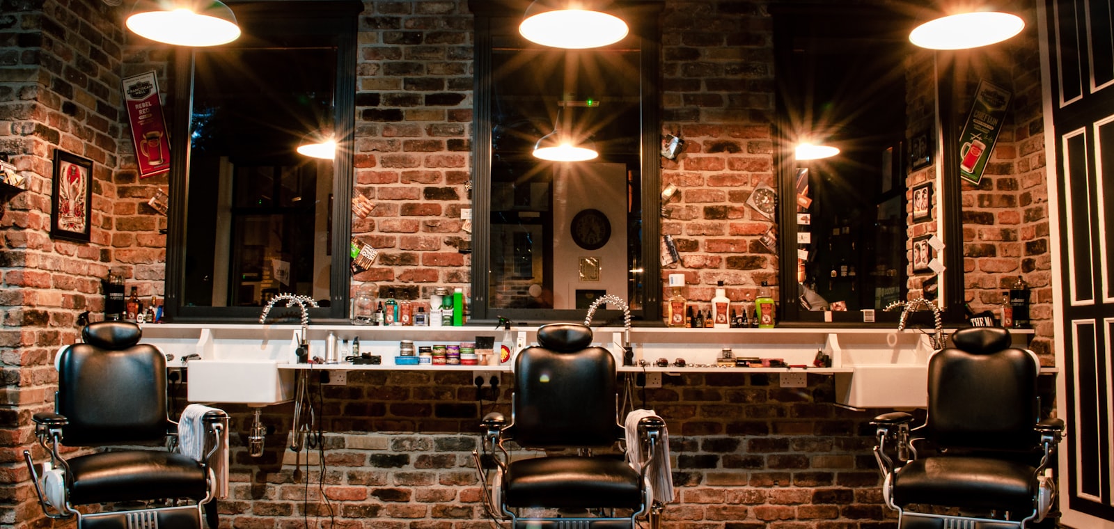 The barbershop interior at night, three chairs lit by pendant lamps against exposed brick